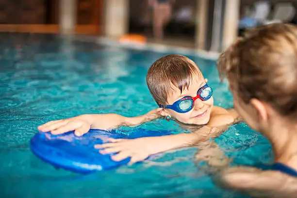 Happy little boy during swimming lesson smiles at the instructor. The boy is aged 6 and is wearing swimming goggles. The boy is using a kickboard.