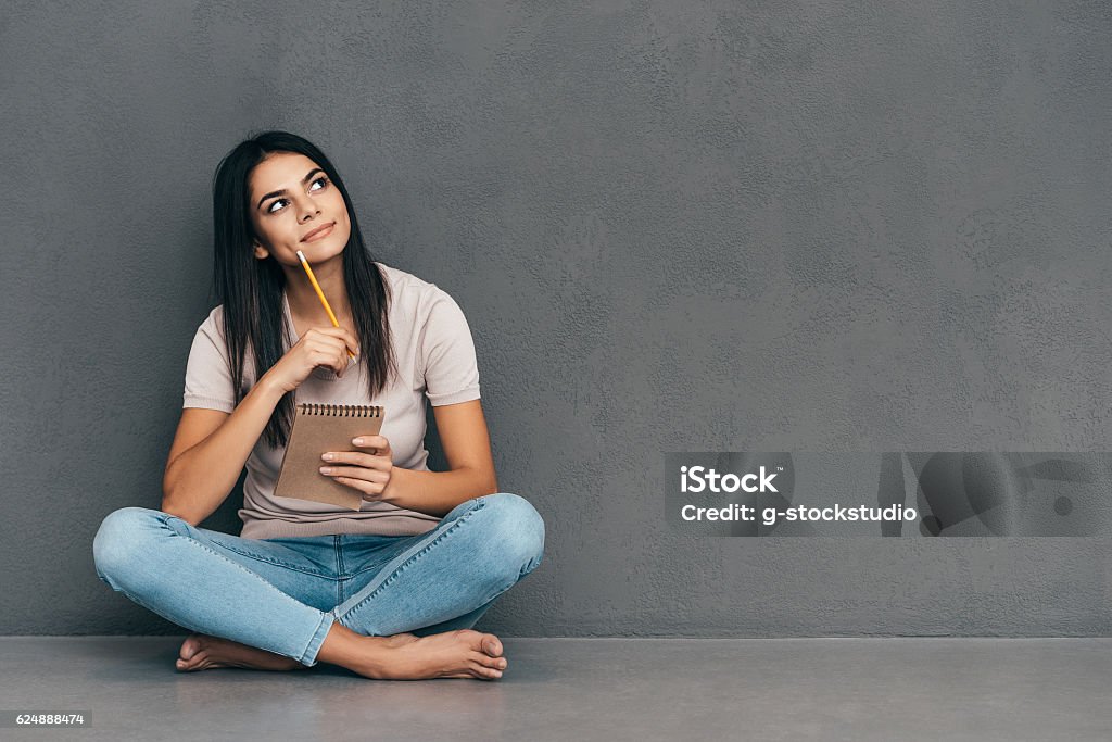Waiting for inspiration. Attractive young woman in casual wear holding notebook and pen while sitting barefoot and against grey background Contemplation Stock Photo
