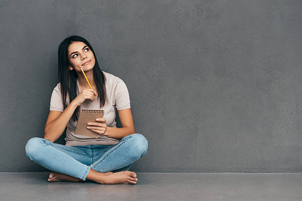 Waiting for inspiration. Attractive young woman in casual wear holding notebook and pen while sitting barefoot and against grey background cross legged stock pictures, royalty-free photos & images