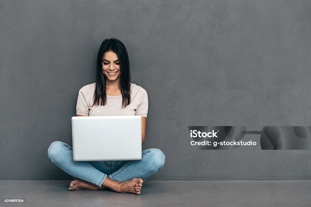 Working on laptop. Attractive young woman in casual wear working on laptop and smiling while sitting barefoot and against grey background Laptop Stock Photo