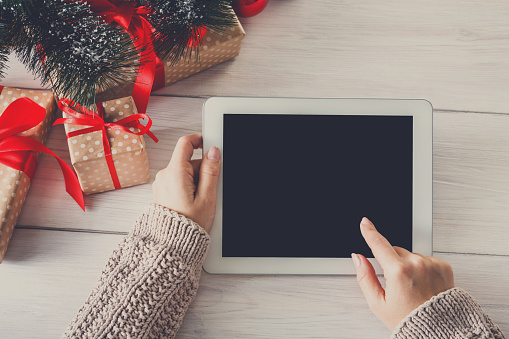 Christmas online shopping above view on wood. Female buyer touch screen of tablet, copy space. Woman has coffee, buys presents near christmas tree, among gift boxes. Winter holidays sales background