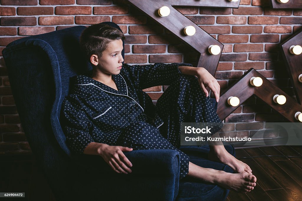 Bellissimo ragazzo adolescente alla moda seduto su una sedia a casa - Foto stock royalty-free di Pigiama