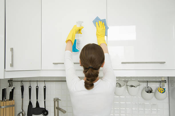 Rear view at young woman cleaning white kitchen wall cabinet Rear view at an attractive young woman cleaning a surface of white kitchen wall cabinet, wearing rubber protective yellow gloves, with rag and spray bottle detergent. Home, housekeeping concept cleaning cupboard stock pictures, royalty-free photos & images