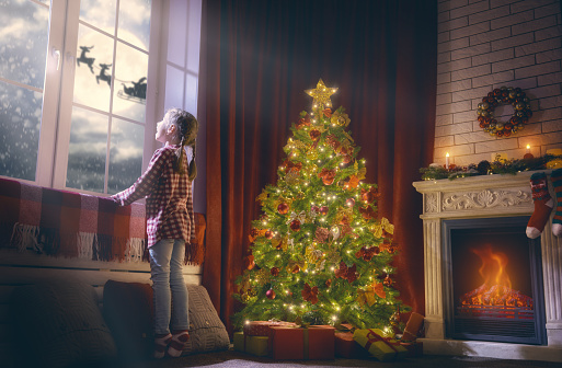 Merry Christmas and happy holidays! Cute little child girl looking by window. Christmas miracle Santa Claus flying in his sleigh against moon sky. Room decorated on Christmas. Kid enjoy the holiday.