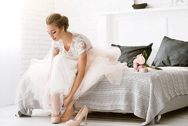 Beautiful bride sitting in the white colored room The last minutes. Charming delighted young bride sitting on the bed in white colored room while wearing her shoes and expressing tenderness wedding shoes stock pictures, royalty-free photos & images