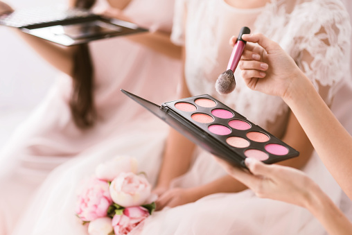Great palette of blusher. Pleasant good looking young bridesmaids sitting in the white bedroom while helping the bride to get ready and holding the palette of blusher