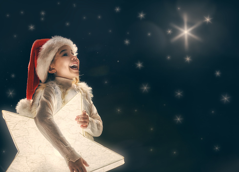 Merry Christmas and happy holidays! Cute little child girl with xmas star. Kid enjoy the holiday. Portrait kid on dark sky background. Christmas legend concept.
