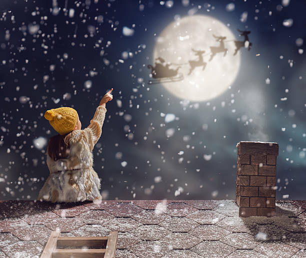 Merry Christmas and happy holidays! Cute little child girl sitting on the roof and looking at Santa Claus flying in his sleigh against moon sky. Kid enjoy the holiday. Christmas legend concept.