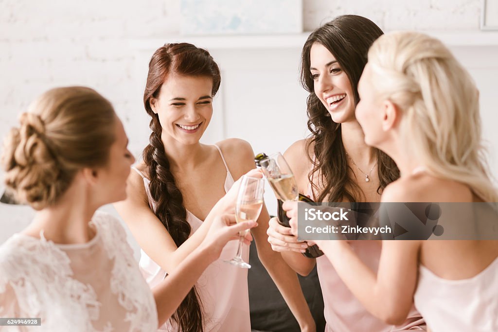 Joyful bride and bridesmaids having party at home We relaxing together. Amused smiling young girls sitting in the white bedroom while having fun and drinking beverage Party - Social Event Stock Photo