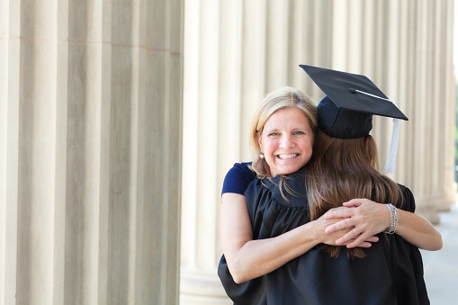 Mother embracing her daughter in a graduation ceremony. The mother is smiling and happy, looking at the camera, posing. A university hall is in the background. Photographed in horizontal format with copy space.