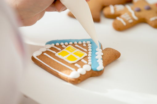 Close up of confectioner hand decorating a gingerbread house, the roof with icing sugar for a snowy effect using a pastry bag. View over a shoulder, lifestyle