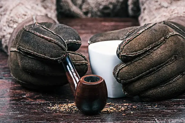 Human hands in winter sheep skin gloves holding cup of coffee and smoking pipe. Front closeup view