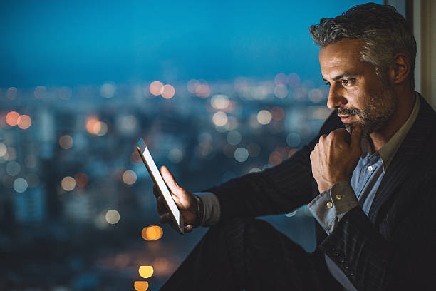 Businessman looking at digital tablet at night Businessman sitting on a windowsill at night and looking at a digital tablet, with cityscape at the background and copy space. street light photos stock pictures, royalty-free photos & images