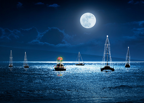 This photo illustration depicts a warm tropical Christmas Holiday scene with a bright full moon, boats decorated with lights and a Palm Tree as a Christmas Tree.