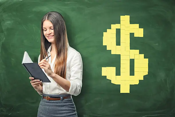A young businesswoman holding her open day planner and writing something in it and yellow sheets of paper forming big dollar sign on background. Daily planning. Time-management. Secretary work.