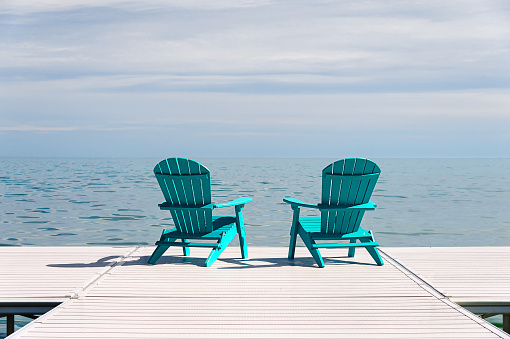 Two turqoise blue muskoka and adirondack chairs on a dock overlooking a blue lake with a sunny day blue sky