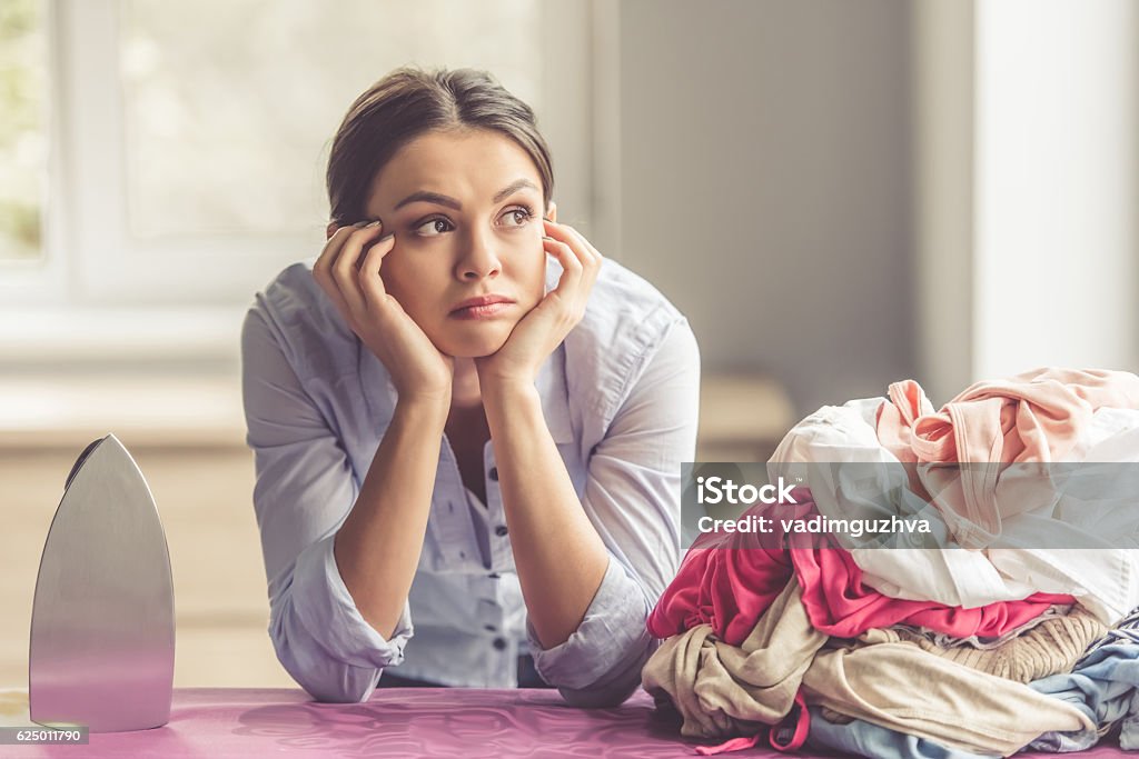 Woman ironing clothes Beautiful bored young woman is leaning on ironing board and looking away while ironing clothes at home Iron - Appliance Stock Photo