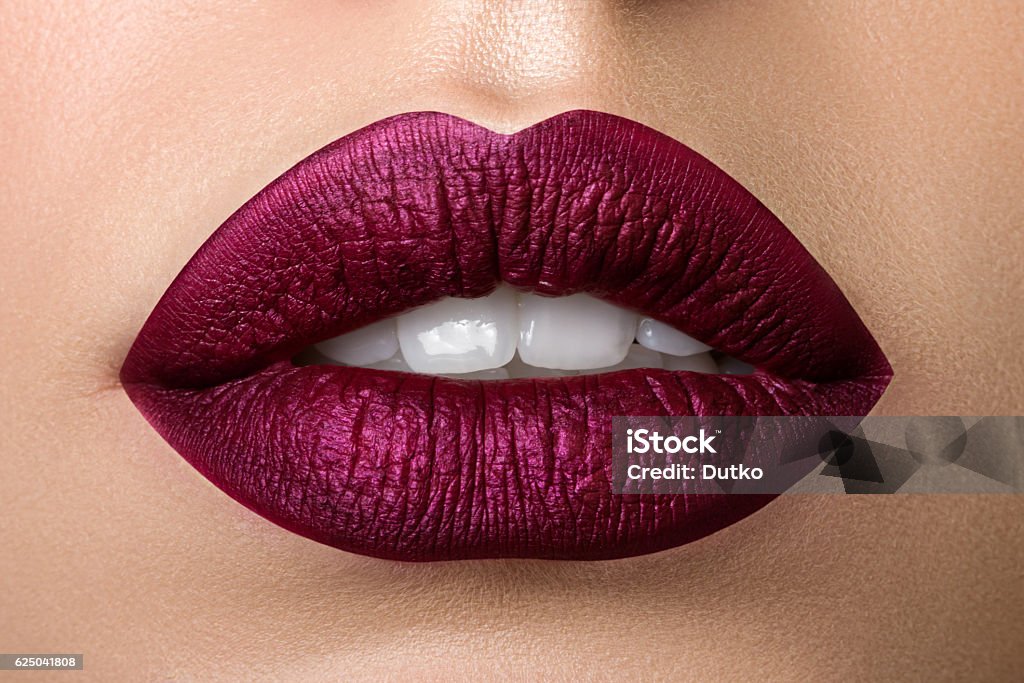 Closeup view of beautiful woman lips with purple matt lipstick Close up view of beautiful woman lips with purple matt lipstick. Open mouth with white teeth. Cosmetology, drugstore or fashion makeup concept. Beauty studio shot. Passionate kiss Human Lips Stock Photo