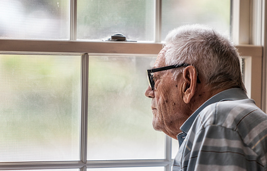 A hearing impaired elderly senior adult man wearing a hearing aid is sitting staring through the hazy, speckled, unwashed window and grungy mesh screen of a living room window at home. He has early stage dementia and will sit motionless for minutes at a time gazing engrossed at the outside world.