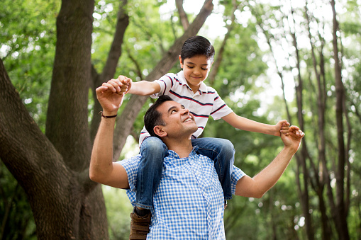 A mature latin father carrying his son on his shoulders, holding hands and smiling at each other in a horizontal medium shot outdoors.
