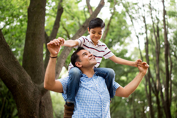 Latin father carrying son on shoulders and looking up A mature latin father carrying his son on his shoulders, holding hands and smiling at each other in a horizontal medium shot outdoors. on shoulders stock pictures, royalty-free photos & images