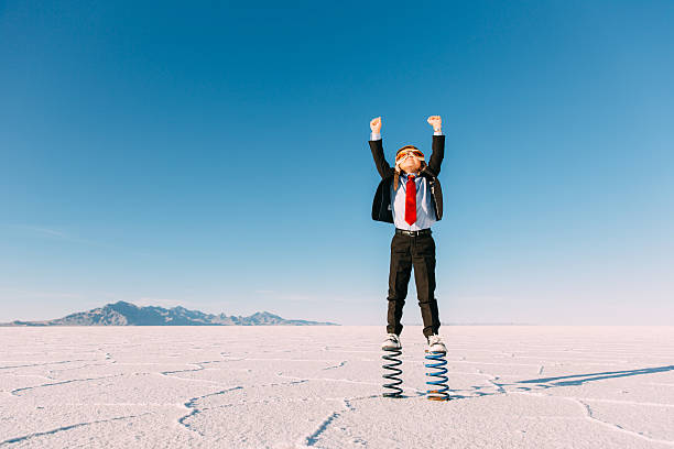 Young Boy Businessman Stands Arms Raised on Springs A young business boy dressed in business suit, flight cap and goggles stands on springs in the Utah desert. He is raising his arms to the sky imagining jumping and flying his business into the sky. Taken at the Bonneville Salt Flats in Utah, USA. taking off activity photos stock pictures, royalty-free photos & images