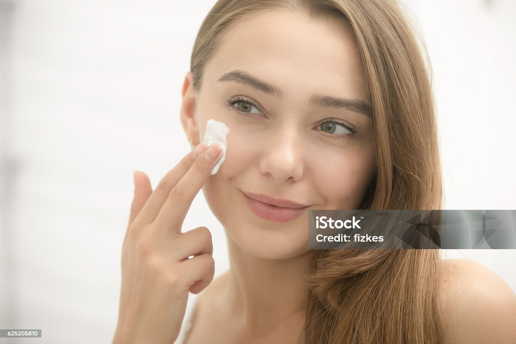 Jeune femme souriante appliquant de la crème sur le visage dans la salle de bain - Photo de Jeunes filles libre de droits