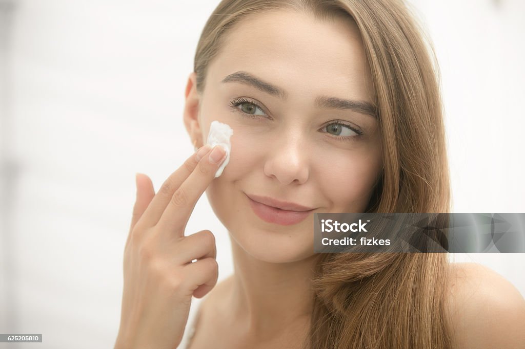 Young smiling woman applying cream to face in the bathroom Young smiling woman applying cream to her face and looking at the mirror at home bathroom. Beauty, skin care concept, lifestyle Teenage Girls Stock Photo