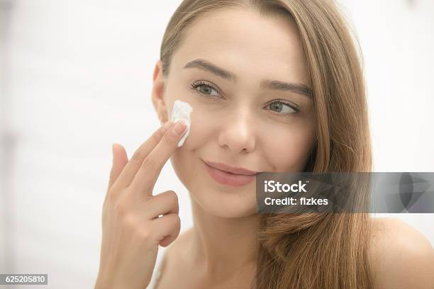 Young Smiling Woman Applying Cream To Face In The Bathroom Stock Photo - Download Image Now