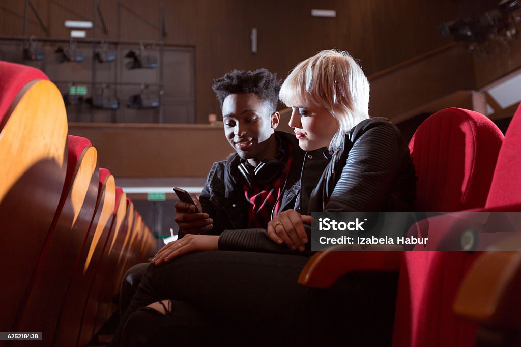 Multi ethnic couple in the movie theater Blonde girl and afro american young guy in the cinema. Side view. Guy using smart phone. Smart Phone Stock Photo