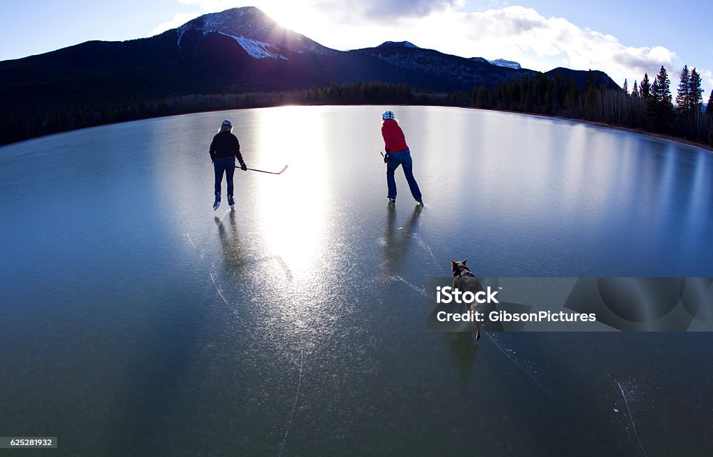Canada Pond Ice Skating Two women enjoy a winter skate on an outdoor pond in the Rocky Mountains of Canada. They are both carrying hockey sticks and are accompanied by their pet dog. Canada Stock Photo