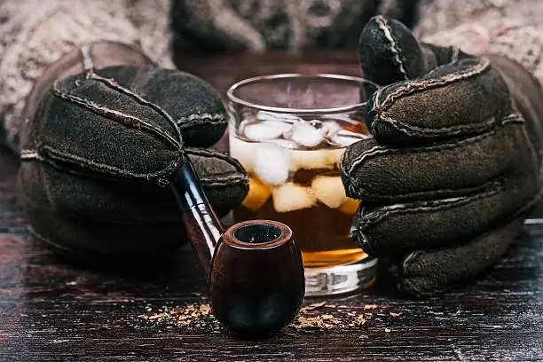 Human hands in winter sheep skin gloves holding glass of wiskey and smoking pipe. Front closeup view
