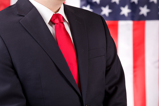 A man in a suit and red tie standing by an American flag.