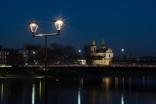 Church in Krakow on river bank. In foreground is a street lantern. In background bridge and a church shot at night