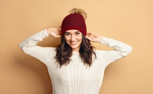 Funny Hipster Girl in Knitted Sweater and Beanie Hat over beige soft Background. Trendy Casual trendy young woman in winter outfit