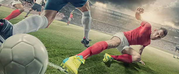A low angle, close up image of a professional soccer on the ground, performing a sliding tackle against an opposition player who is jumping over.  The action is taking place on an outdoor soccer pitch in a generic floodlit football stadium full of spectators under a dramatic, stormy evening sky.