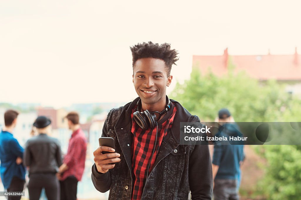Afro american young guy smiling outdoor Outdoor portrait of afro american young guy smiling at camera, holding smart phone in hand. Group of people in the background. Youth Culture Stock Photo