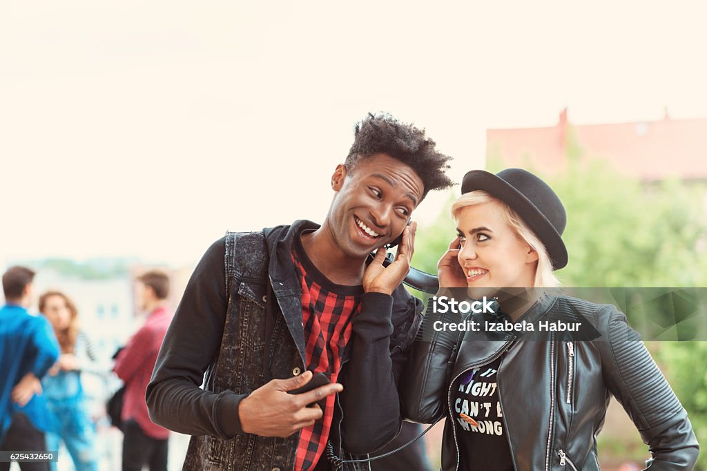 Funky young couple listening to music outdoor Outdoor portrait of funky young couple - afro amercian cool guy and beautiful blonde woman, listening to the music together. Group of people in the background. 20-29 Years Stock Photo