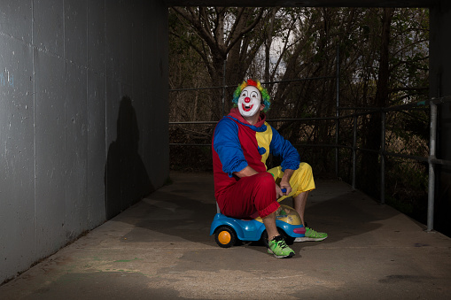 A man in a clown suit rides a small children's ride on car under a bridge