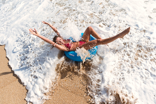 young woman at the shoreline of Porthcurno Beach, South Cornwall on a bright Sunny day, sat in a blue inflatable rubber ring, enjoying the beach.