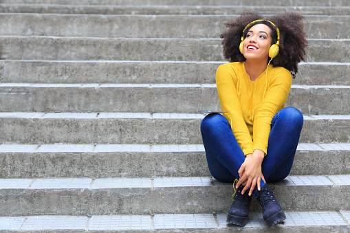 Attractive young woman sitting on stairs and listening music. Yellow headphones on head, wears yellow blouse and blue jeans, with crossed legs. Looking up. Stone stairs on background.