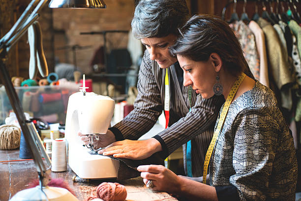 seamstress teaching her assistant how to use the sewing machine - sewing tailor sewing machine women imagens e fotografias de stock