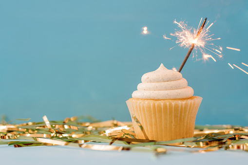 A cupcake sits on a table against a blue background during a New Year's Eve party. This white cupcake is a sweet new year's dessert, and it is surrounded by tinsel, glitter, and has a sparkler on it.