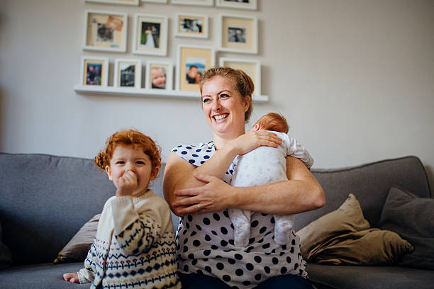 Happy Little Family Mother with her two children in the living room of their home. The mother is holding a baby and the other little girl is laughing and looking at the camera while sucking her thumb. 2 3 years photos stock pictures, royalty-free photos & images