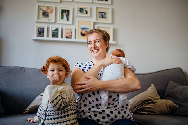 Happy Little Family Mother with her two children in the living room of their home. The mother is holding a baby and the other little girl is laughing and looking at the camera while sucking her thumb. mom and baby eye contact stock pictures, royalty-free photos & images