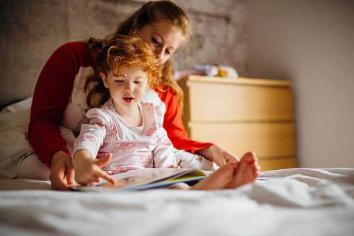 Little girl reading a bedtime story with her Mother in her bed. They are sitting together and the little girl is pointing at one of the pictures.