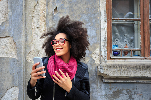 Beautiful young woman making selfie in front of vintage building. Wears black jacket and pink pullover, with red eyeglasses.