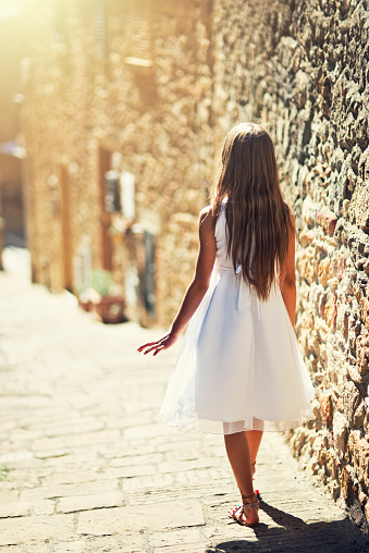 Teenage girl or a young woman walking sunny narrow street of a charming italian town. The girl is wearing a white dress.