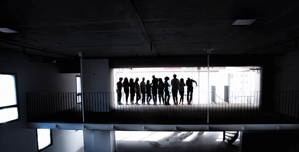 The silhouette of a group of 12 people against a back lit window in a dark warehouse.