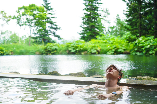 Front view of young man with white towel on his head while lying and enjoying hot mineral water in outdoor onsen (hot spring) near Niseko on Hokkaido. In background are trees and grey sky of cloudy and rainy weather.  This is traditional way of having bath in Japan.